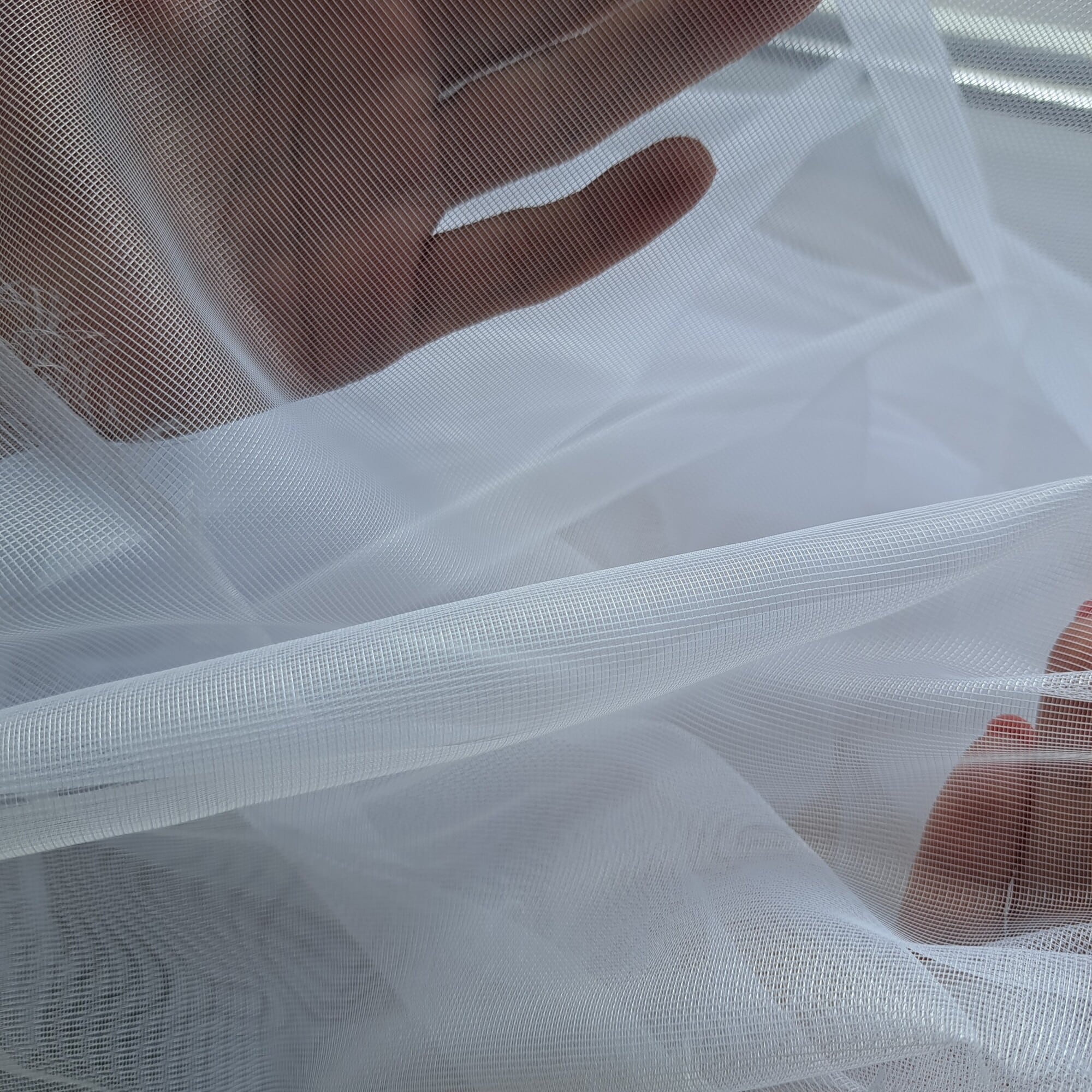 Dłoń unosząca przezroczystą, białą tkaninę w świetle dziennym, delikatna tekstura materiału.