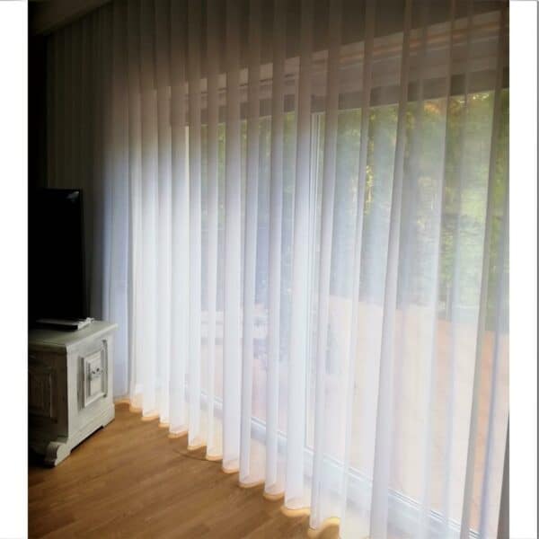 Białe zasłony wiszące na dużym oknie, przepuszczające naturalne światło do jasnego wnętrza pokoju.