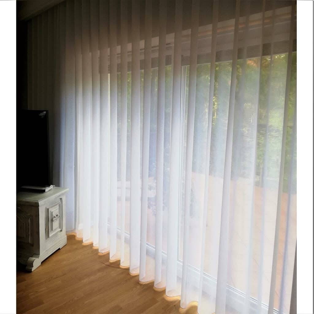 Białe zasłony wiszące na dużym oknie, przepuszczające naturalne światło do jasnego wnętrza pokoju.