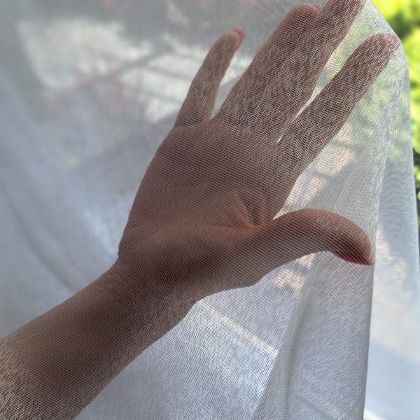 Dłoń za przezroczystą firanką z widokiem na zewnątrz, delikatnie oświetlona słońcem.