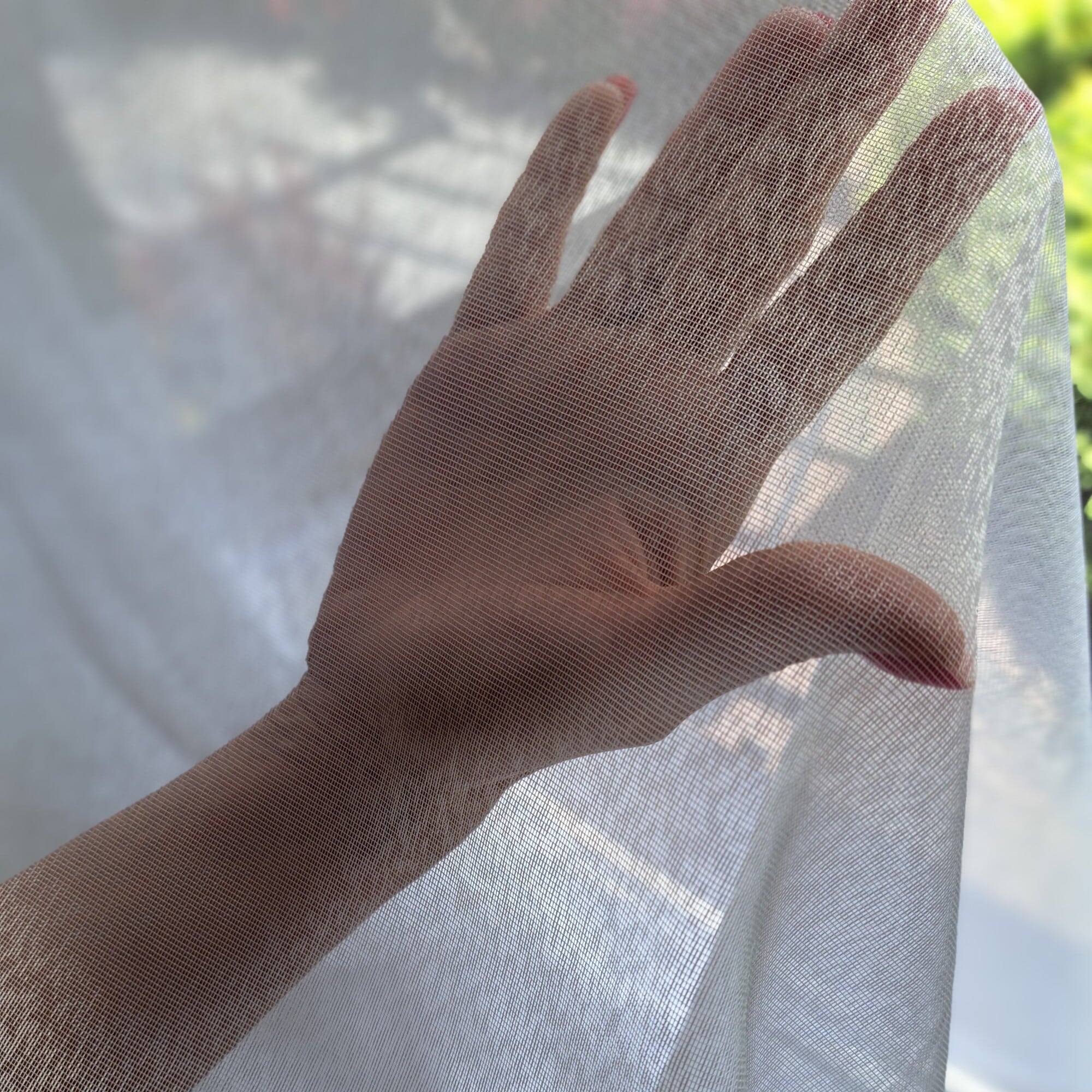 1211 - 1 Dłoń za przezroczystą firanką z widokiem na zewnątrz, delikatnie oświetlona słońcem.