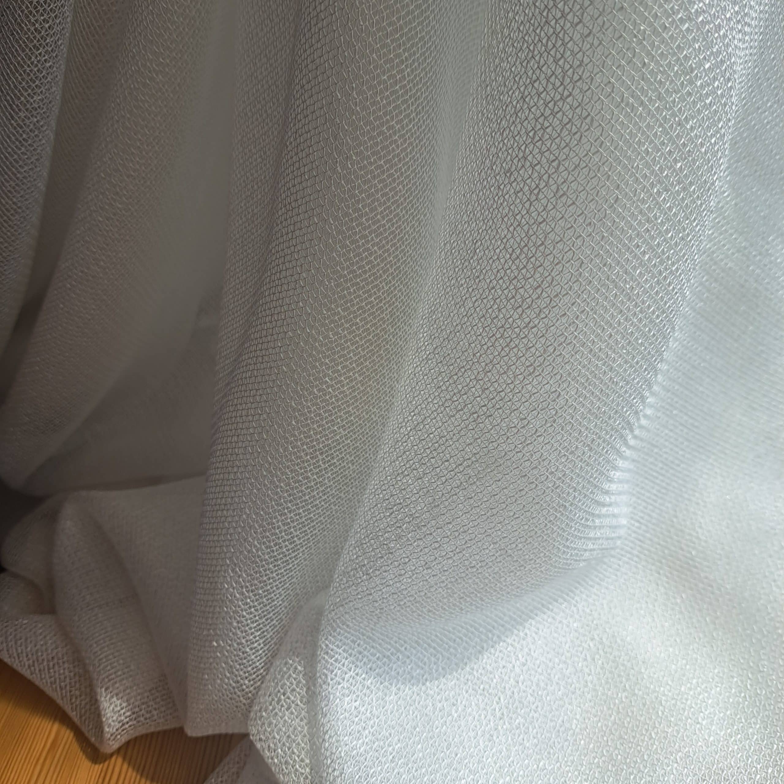 302 bi 4 Biała tiulowa zasłona w delikatnym świetle, z widocznym teksturem materiału na tle drewnianej podłogi.