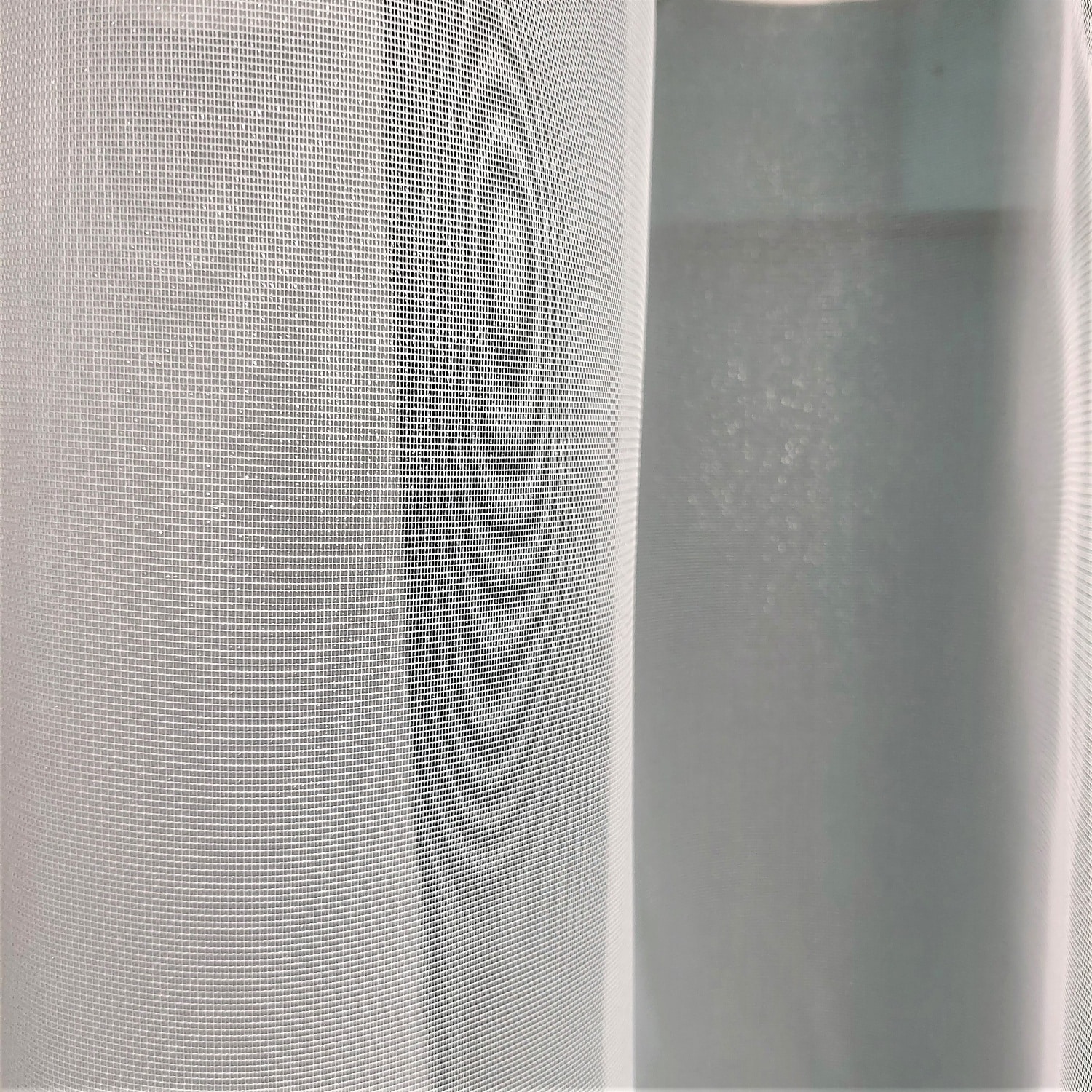 Biała zasłona z delikatnej siatki zawieszona w oknie, przepuszczająca światło dzienne.