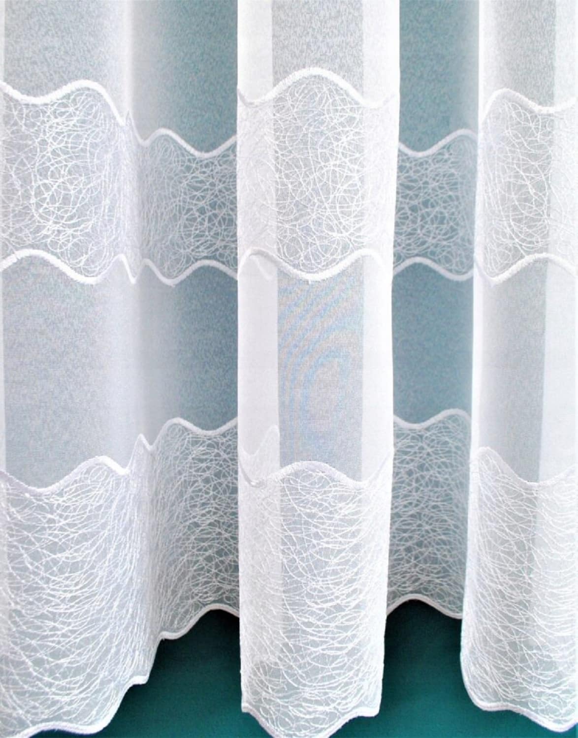107-haft Biała zasłona z ozdobnym wzorem fal, zawieszona na tle niebieskiej ściany, tworząca elegancki efekt w wnętrzu.