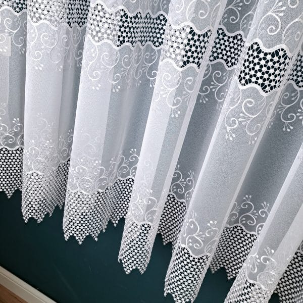 Białe firany z delikatnym wzorem i ażurowymi wstawkami, wiszące na tle zielonej ściany.