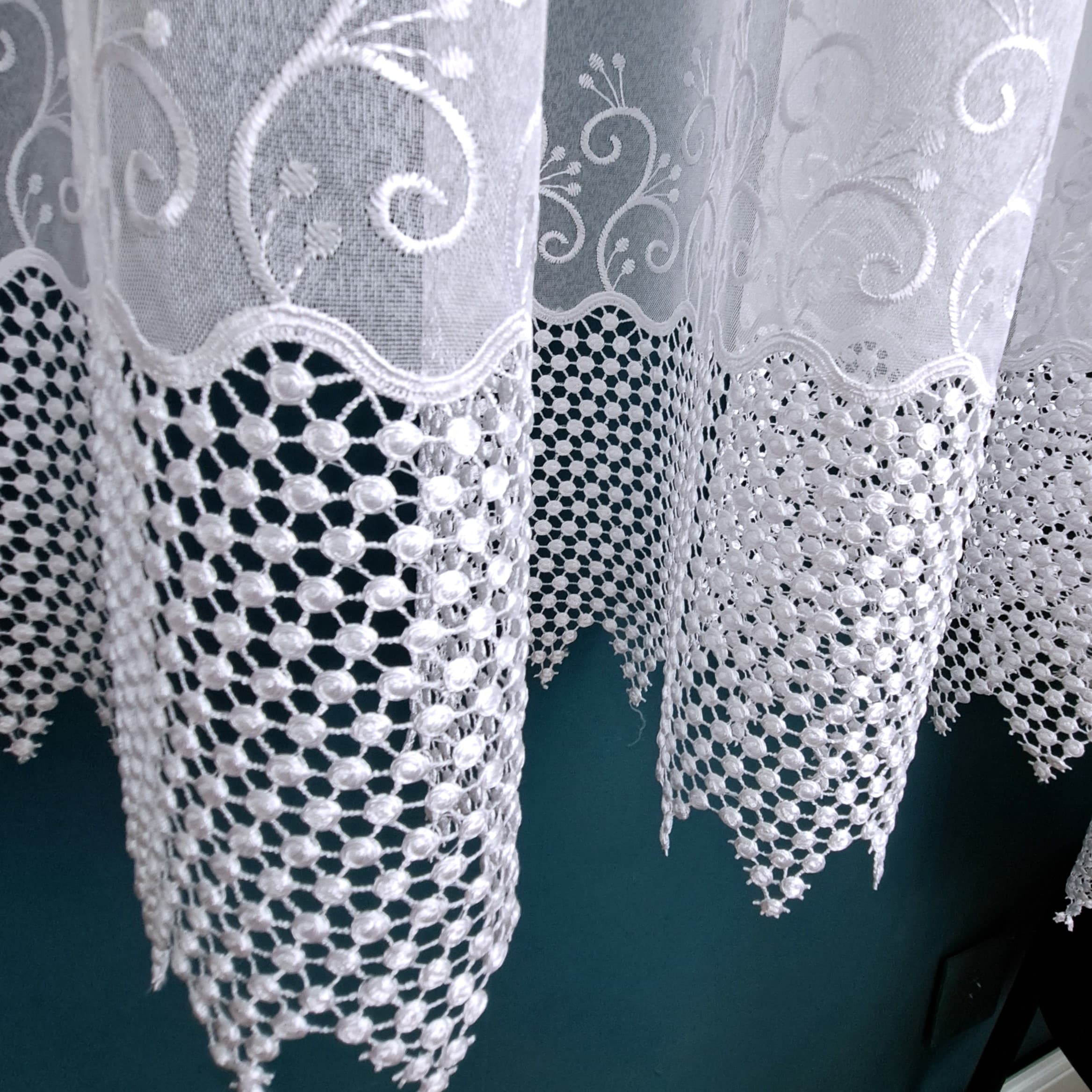 Koronkowa firanka z delikatnym wzorem na tle ciemnej ściany, elegancka dekoracja wnętrza.