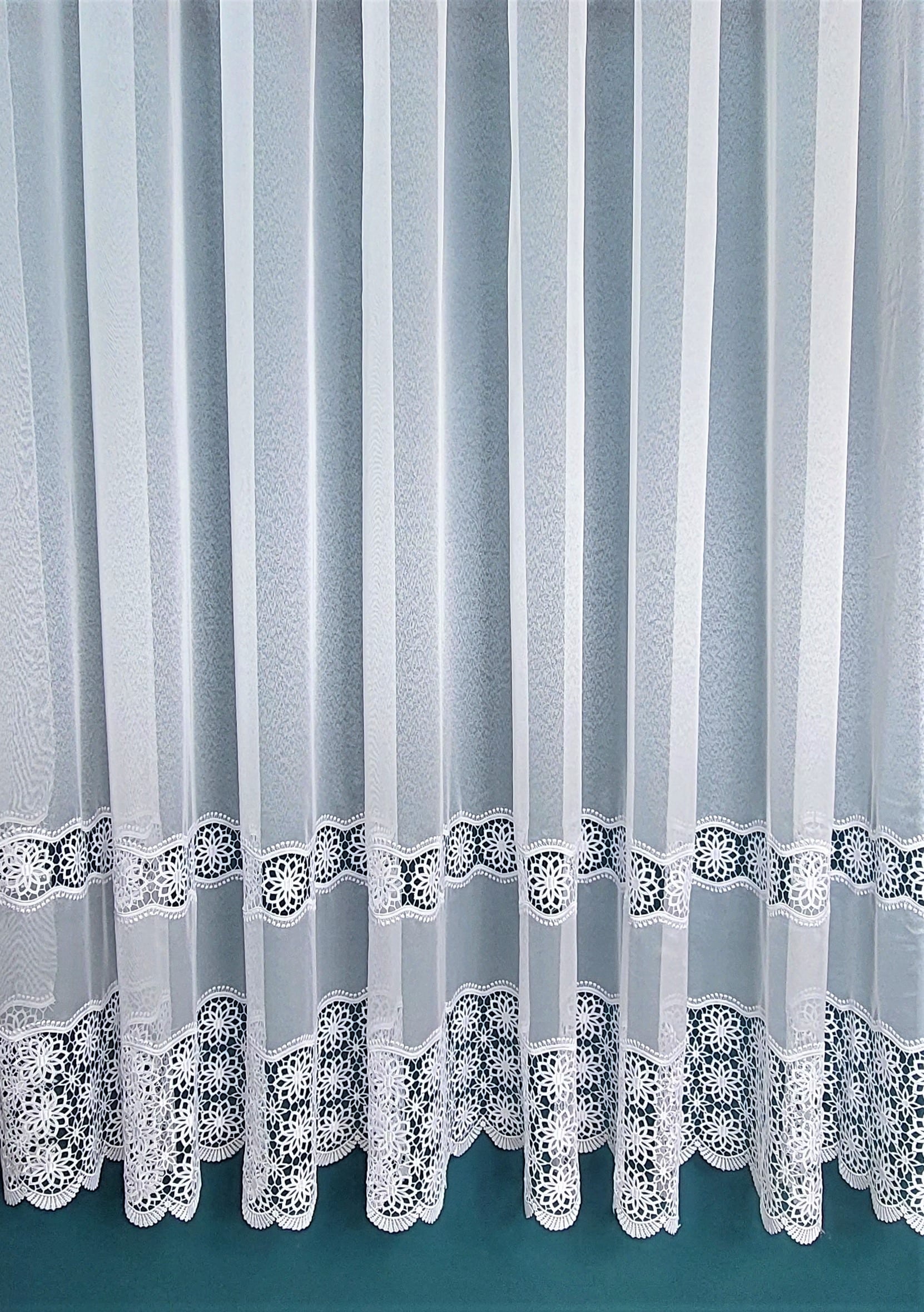 Białe firany z koronkowym wzorem na dole, delikatny materiał zapewnia elegancki wygląd i przepuszcza światło.