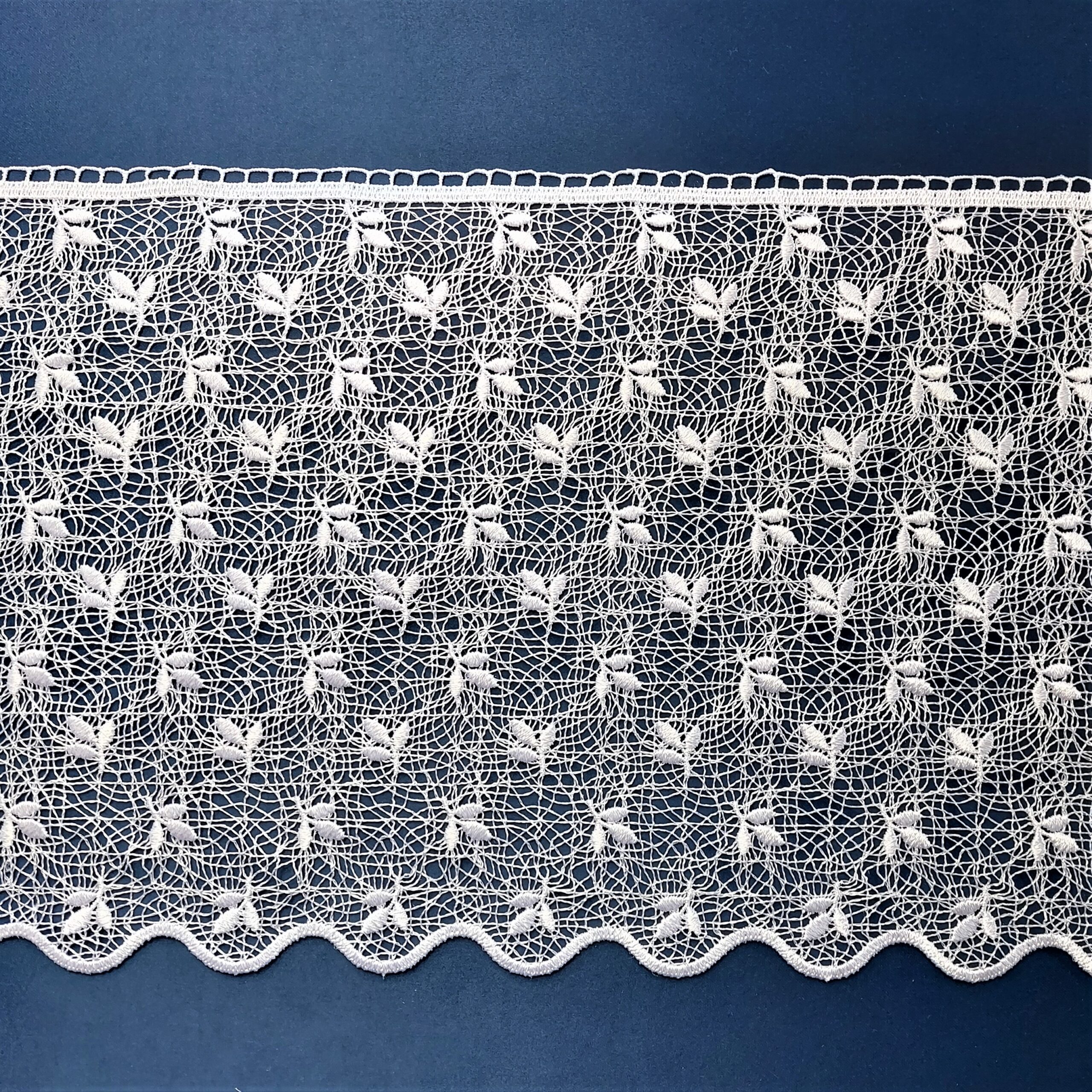 Koronkowy materiał w białym kolorze na niebieskim tle, ozdobiony wzorem liści i falistym brzegiem.