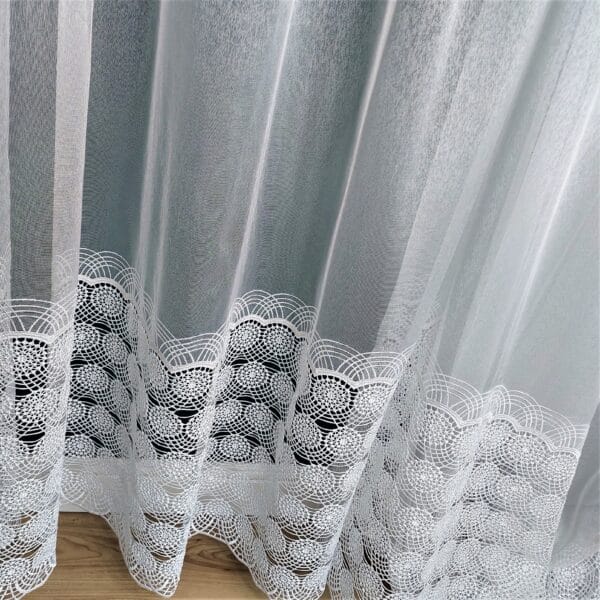 Białe firanki z delikatnym wzorem koronkowym, wiszące przy oknie, zapewniające elegancję i intymność wnętrza.