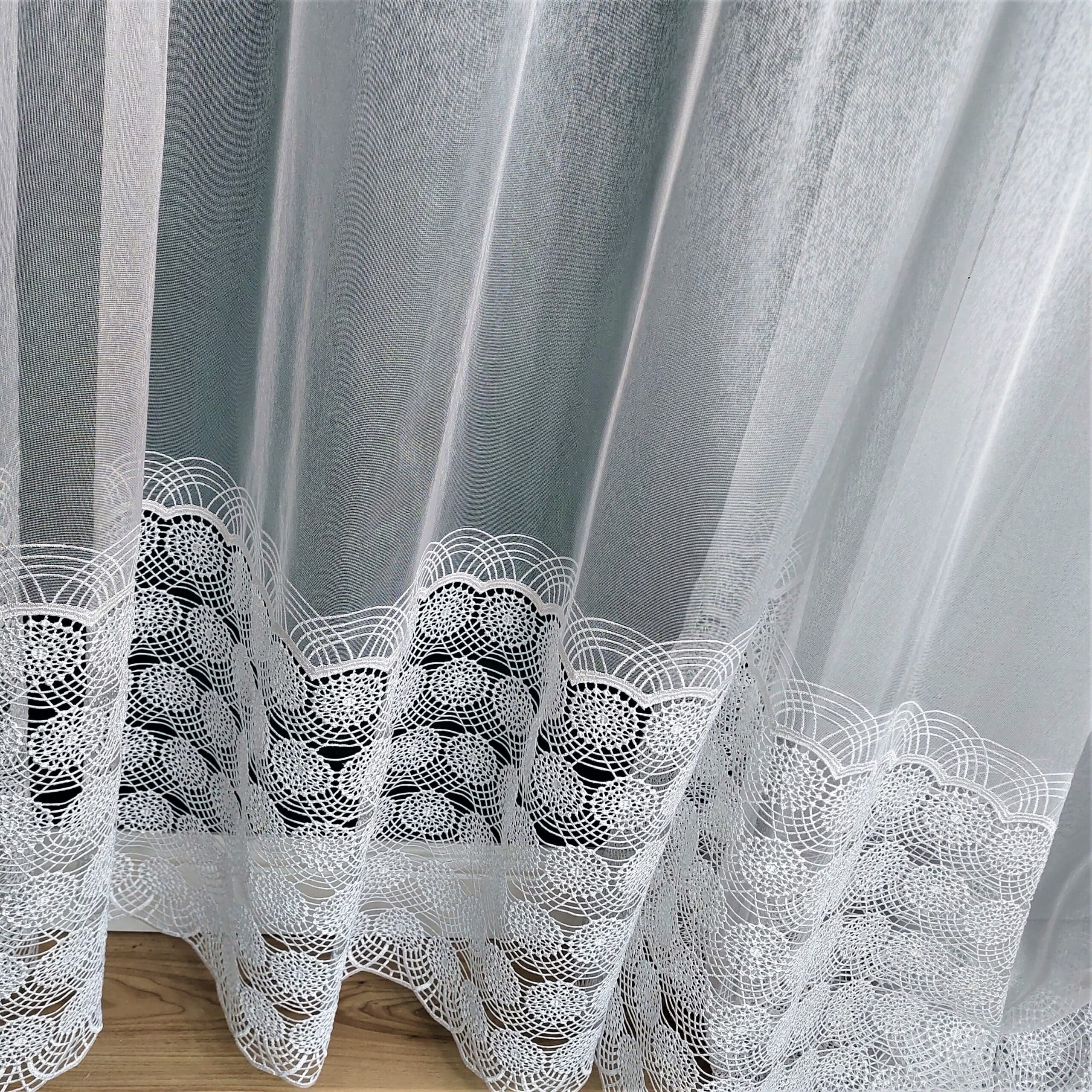 Białe firanki z delikatnym wzorem koronkowym, wiszące przy oknie, zapewniające elegancję i intymność wnętrza.