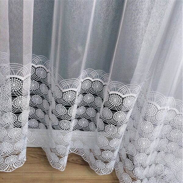 Biała koronkowa zasłona na drewnianej podłodze, ozdobiona delikatnym wzorem na dolnym końcu.