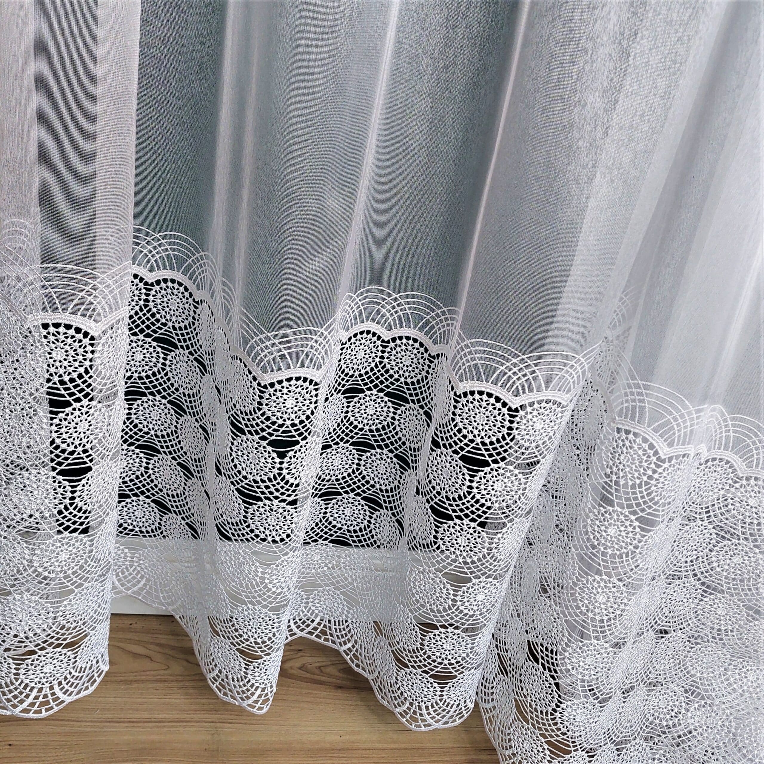 Biała koronkowa zasłona na drewnianej podłodze, ozdobiona delikatnym wzorem na dolnym końcu.