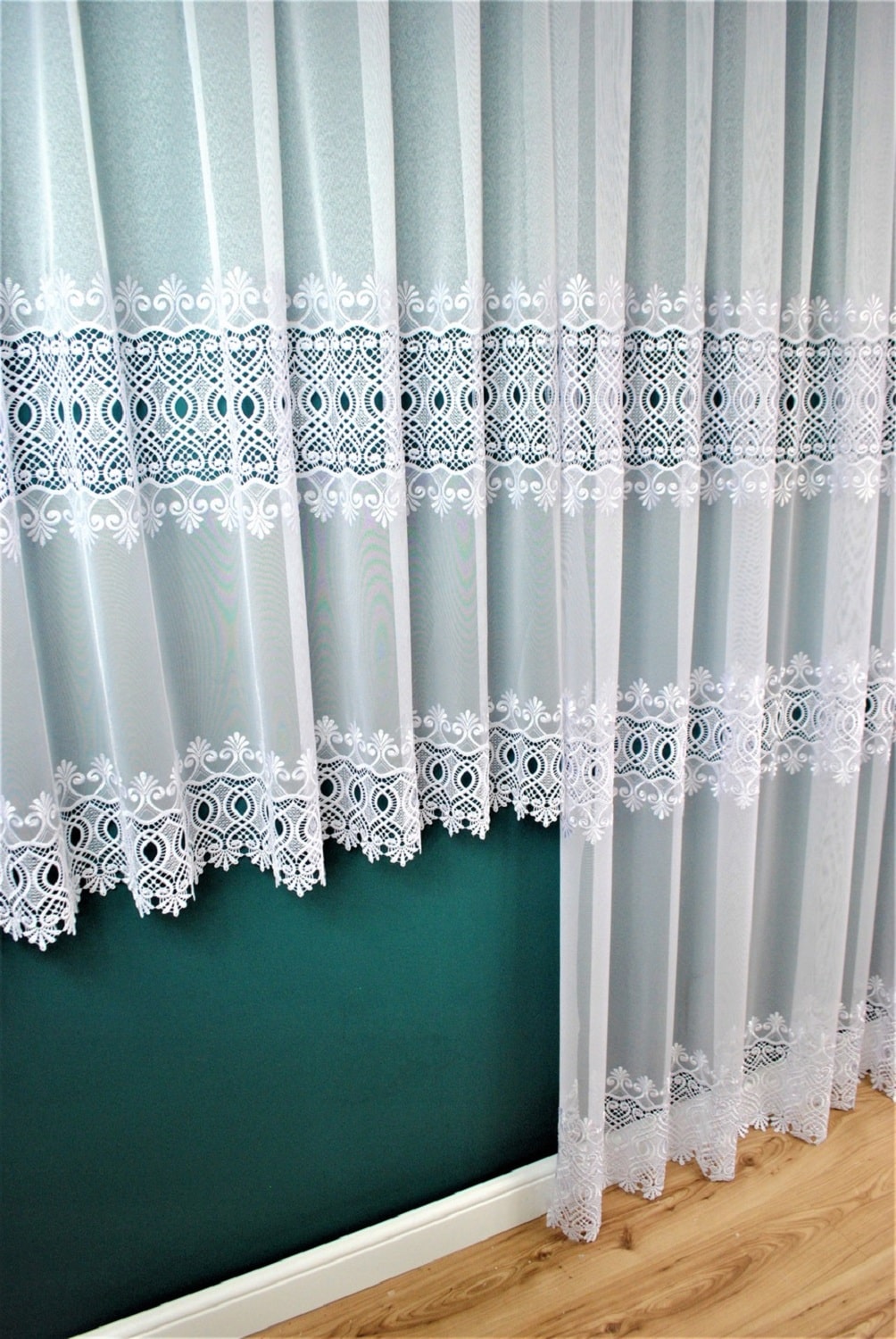 Białe koronkowe firanki w salonie, ozdobne wzory, zielona ściana w tle, drewniana podłoga.
