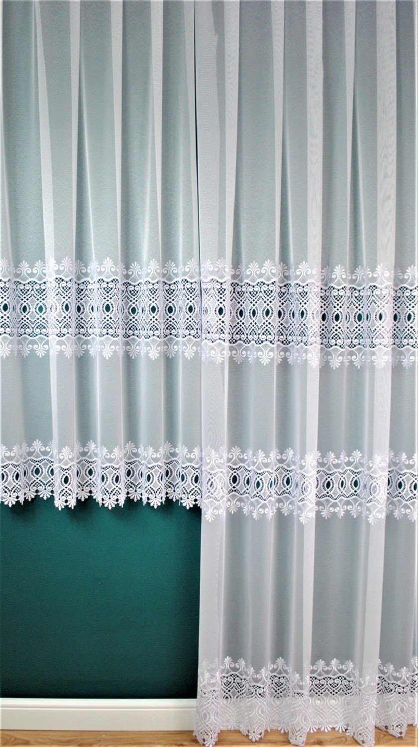 Białe firany z delikatnym wzorem na tle zielonej ściany, drewniana podłoga na dole.