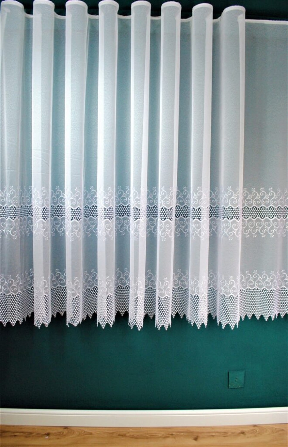 Biała firanka z koronkowym wzorem na tle zielonej ściany, podłoga z jasnego drewna.