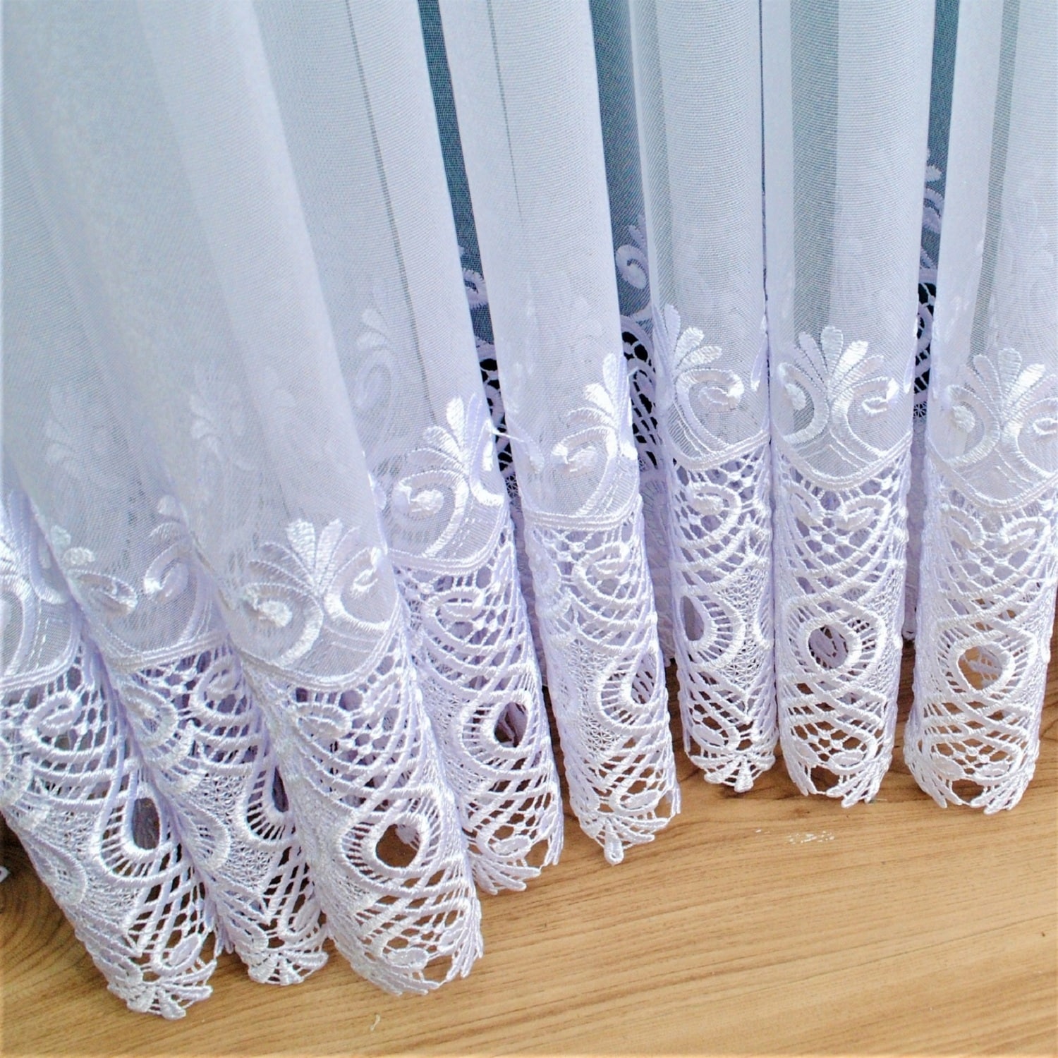 Przezroczysta biała firanka z delikatnym haftem o kwiatowym wzorze na drewnianej podłodze.