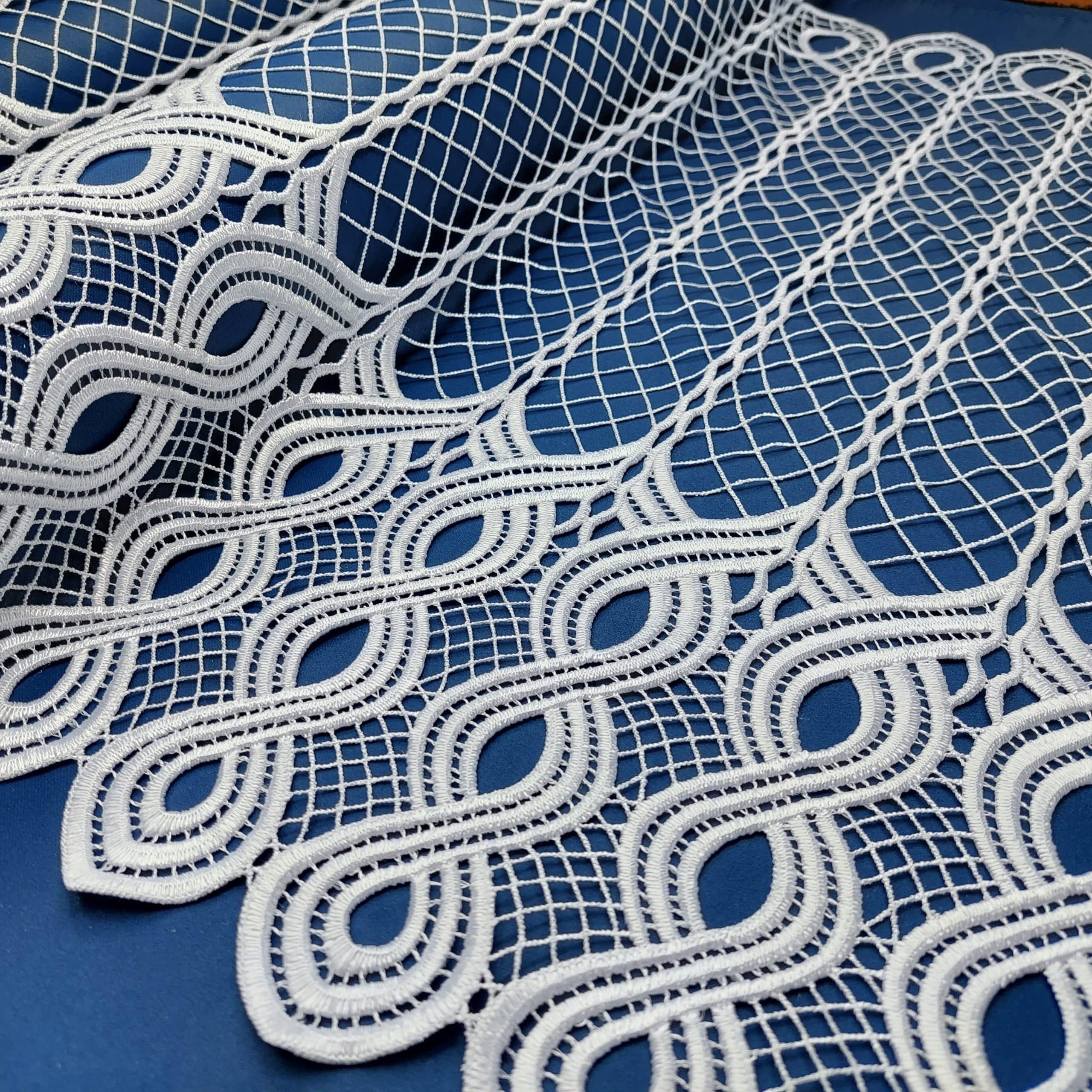 Biała koronka na niebieskim tle, przedstawiająca faliste wzory i siatkę geometryczną. Idealna do dekoracji wnętrz.