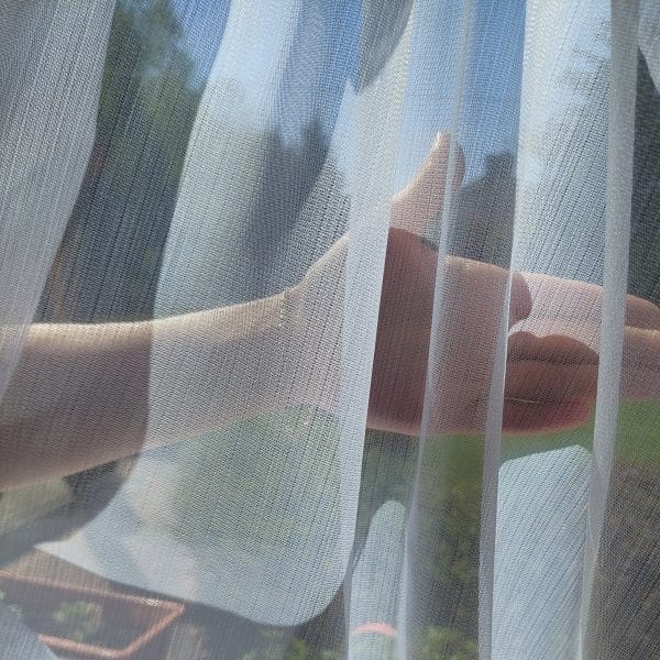 Dłoń za delikatną firanką oświetloną słońcem, tworząca cienie na tkaninie.