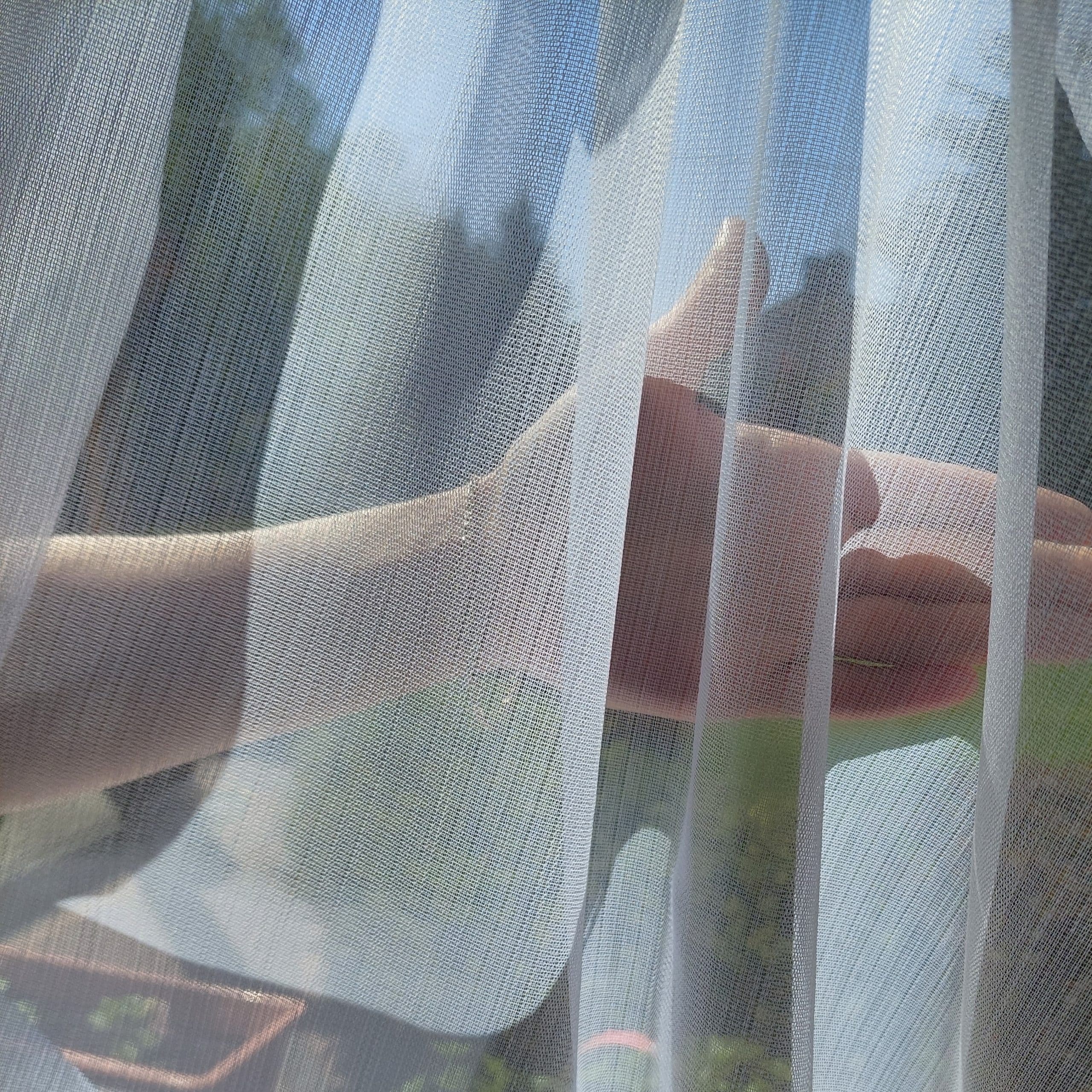 Dłoń za delikatną firanką oświetloną słońcem, tworząca cienie na tkaninie.