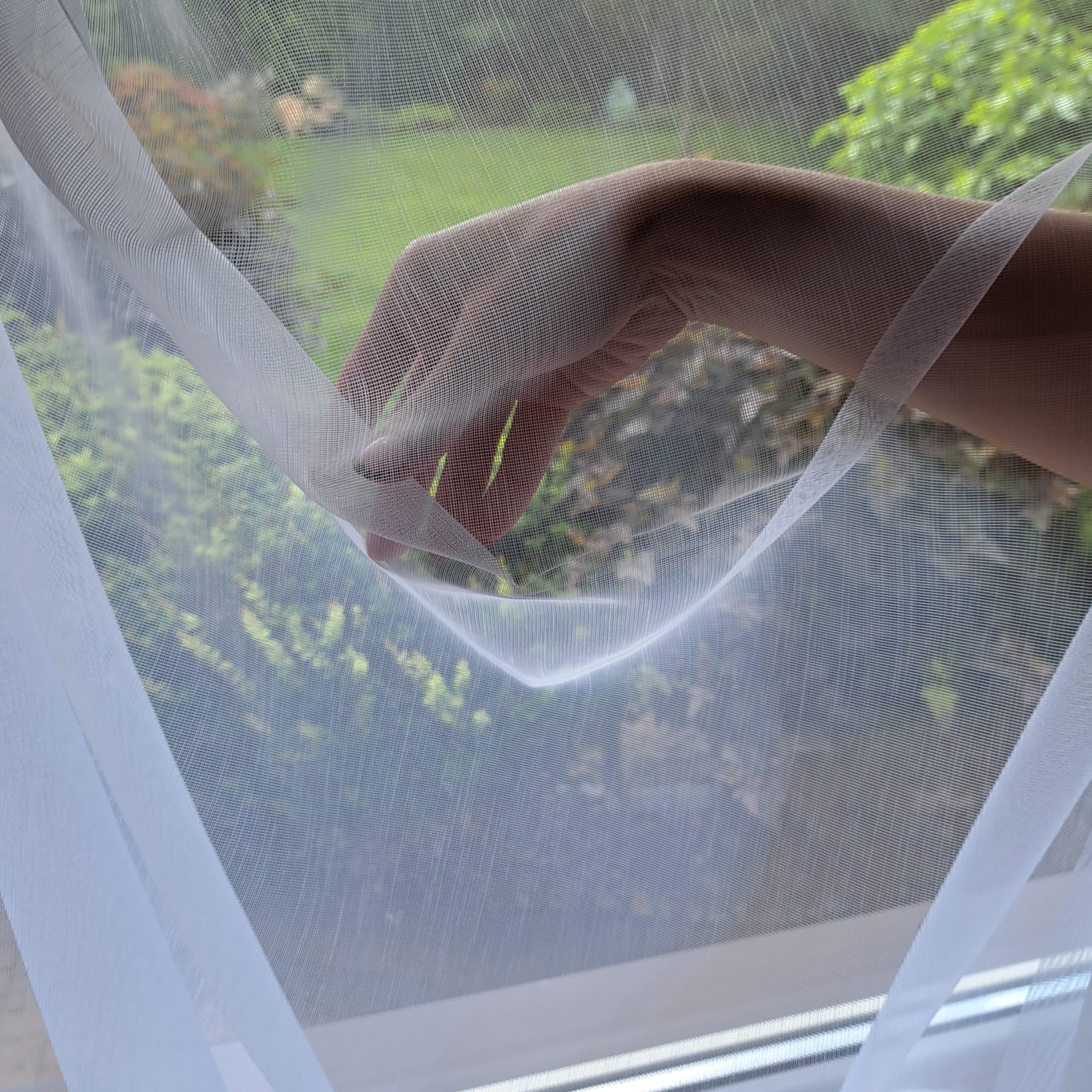 Ręka dotykająca delikatną, białą firankę z widokiem na ogród za oknem.