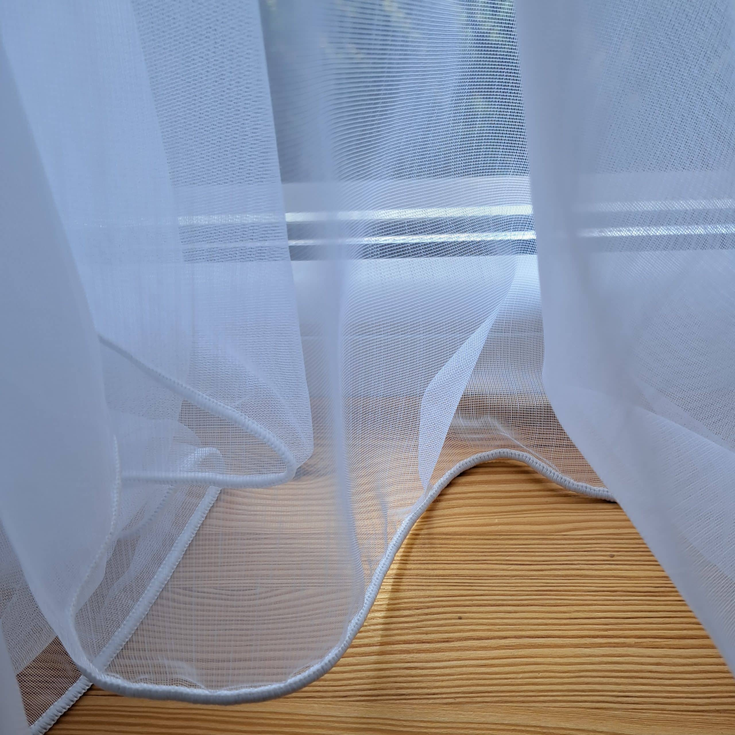 Przezroczysta zasłona unosząca się przy oknie, przepuszczająca światło na jasną drewnianą podłogę.