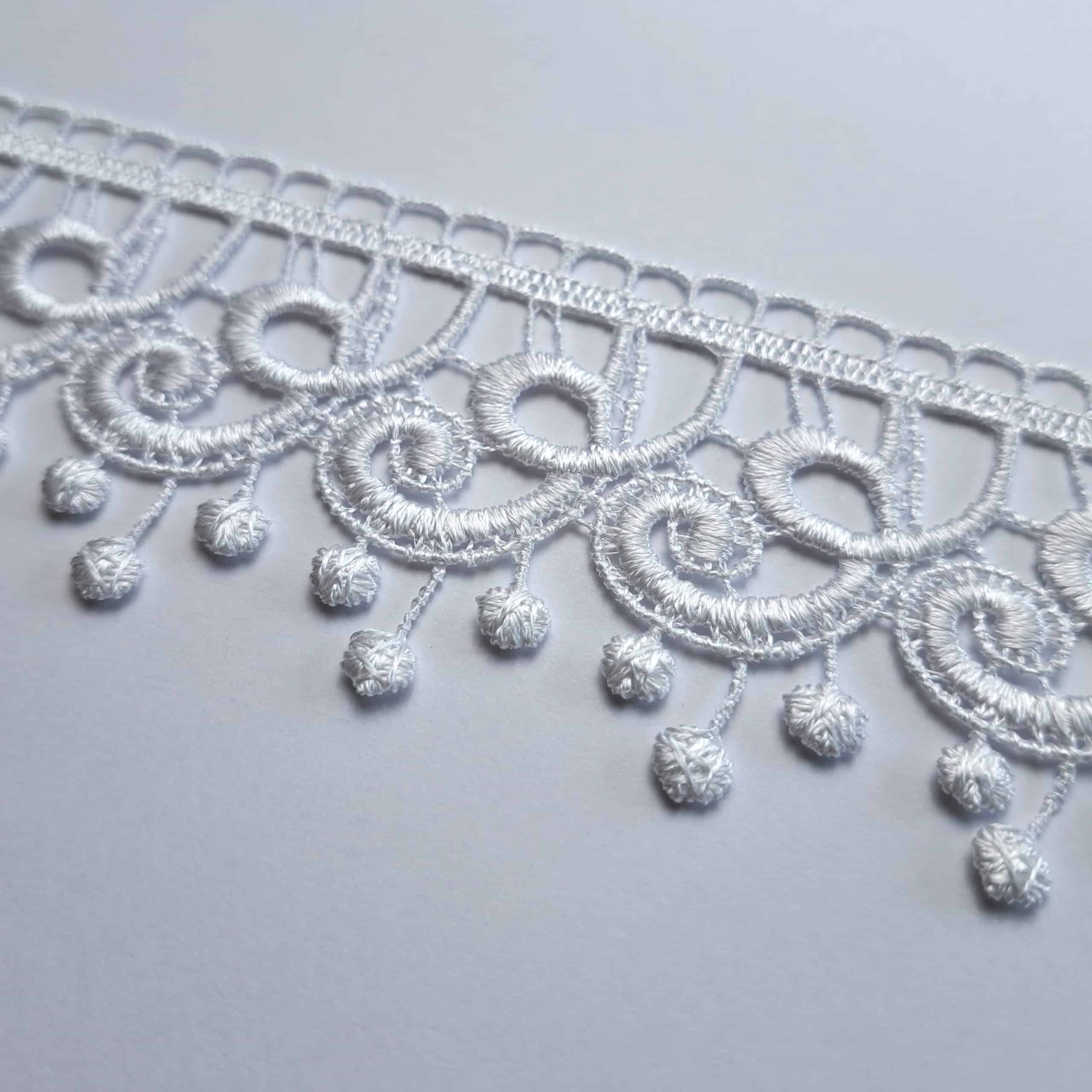 Koronkowy wzór białej tkaniny z ozdobnymi frędzlami na szarym tle.