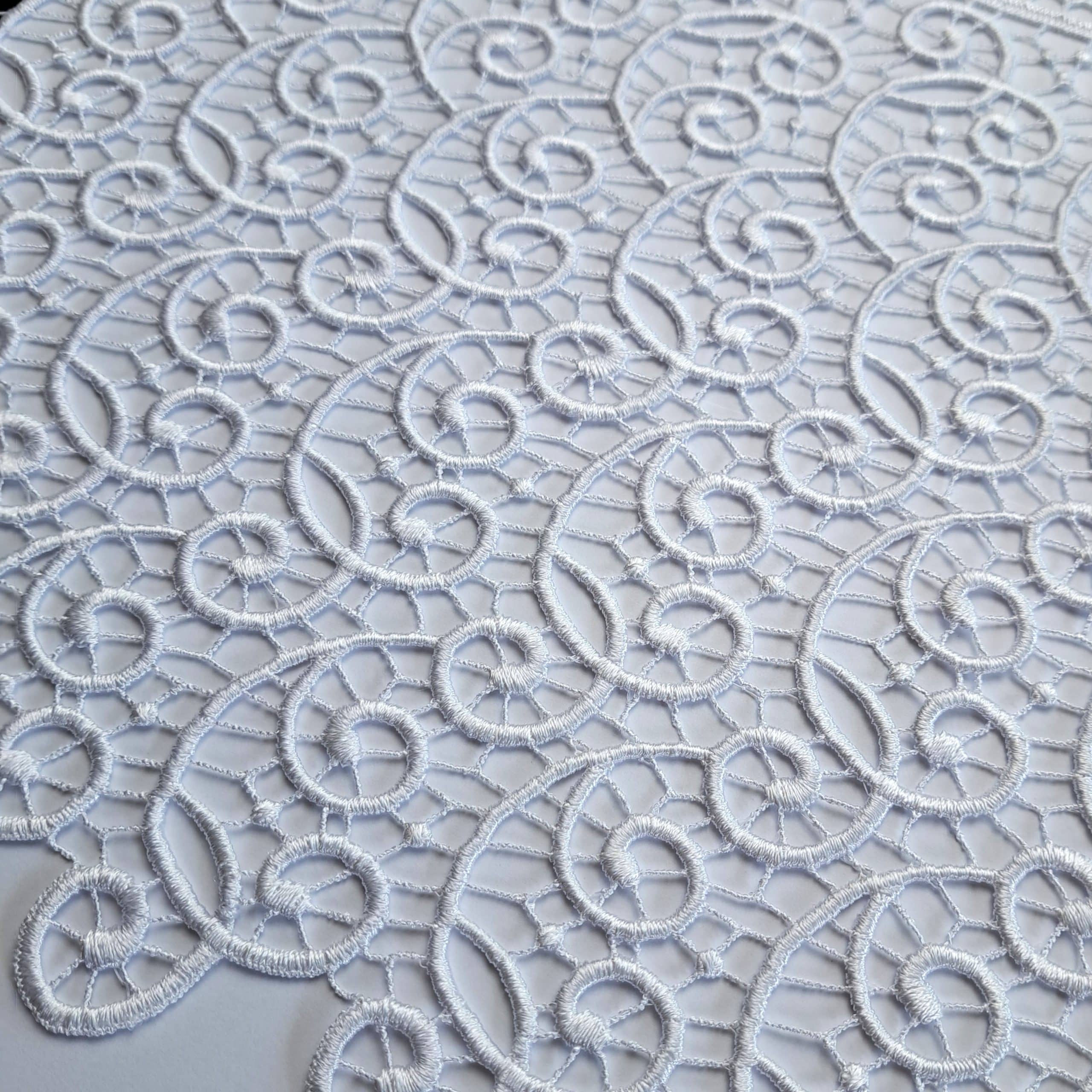 Białe koronkowe zdobienie na tkaninie, wzór złożony z falistych i okrągłych kształtów.
