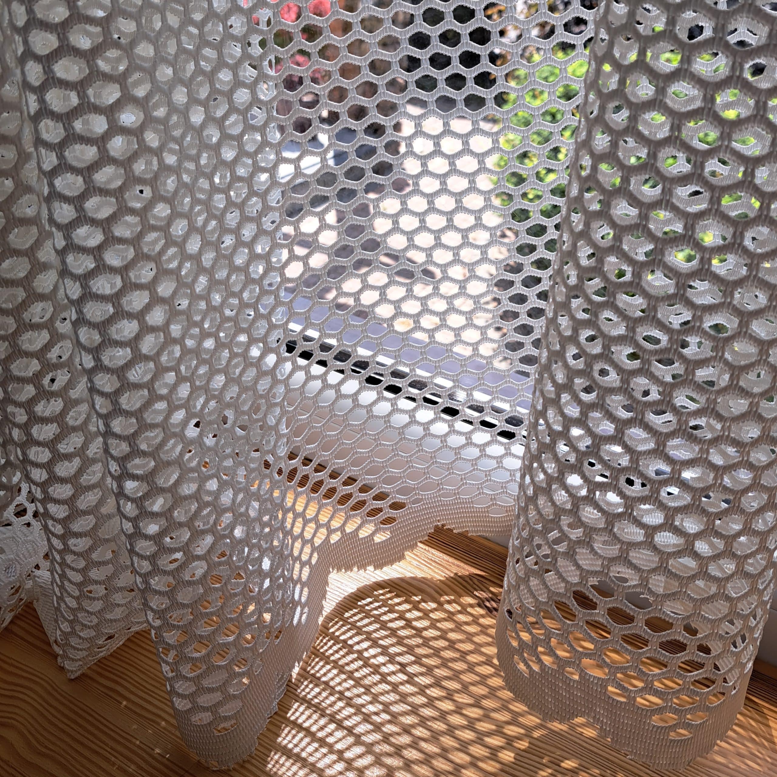 Białe firanki z ażurowym wzorem, światło słoneczne tworzy wzory na drewnianej podłodze za oknem.