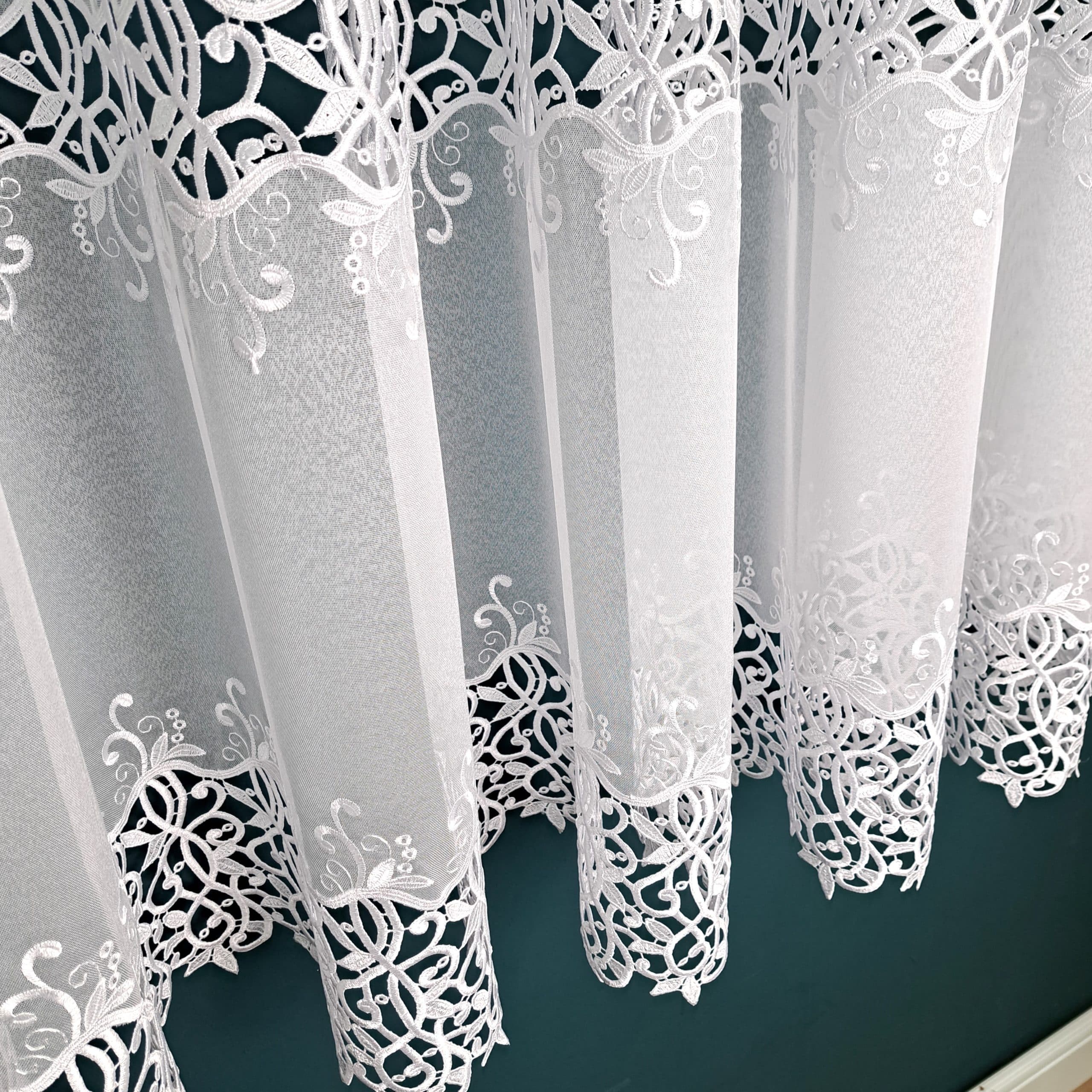 Białe firany z delikatnym, koronkowym wzorem wiszące w fałdach.