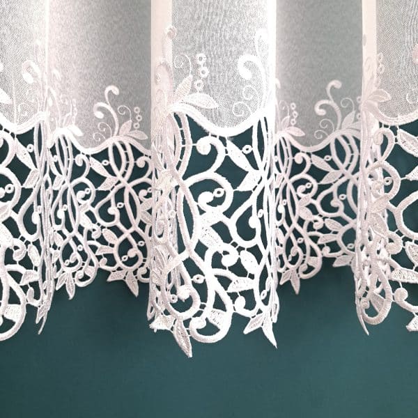 Koronkowa firanka z delikatnym, białym wzorem na tle turkusowej ściany. Elegantna dekoracja wnętrza.