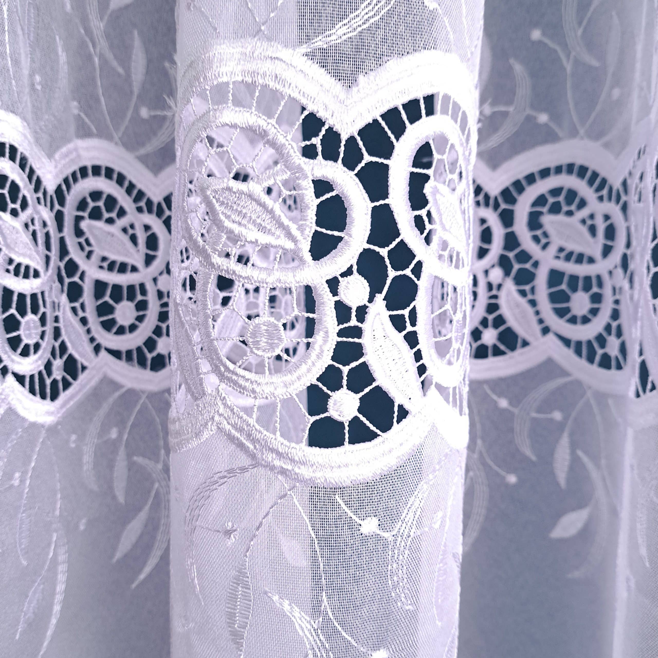 Koronkowa zasłona z delikatnym białym wzorem liści i kwiatów, wpuszczająca światło do wnętrza.