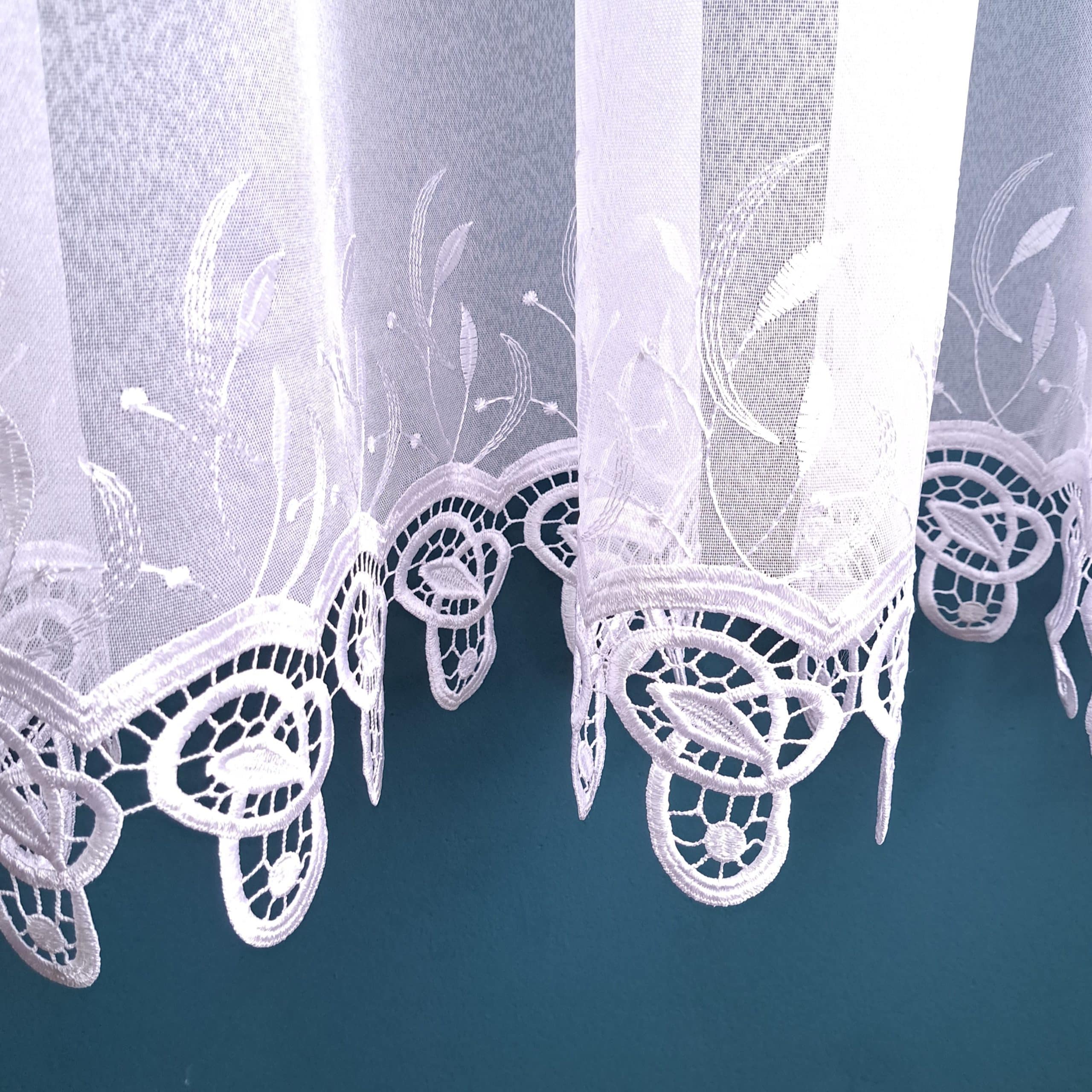 Koronkowa biała firanka z delikatnym haftem na tle turkusowej ściany.